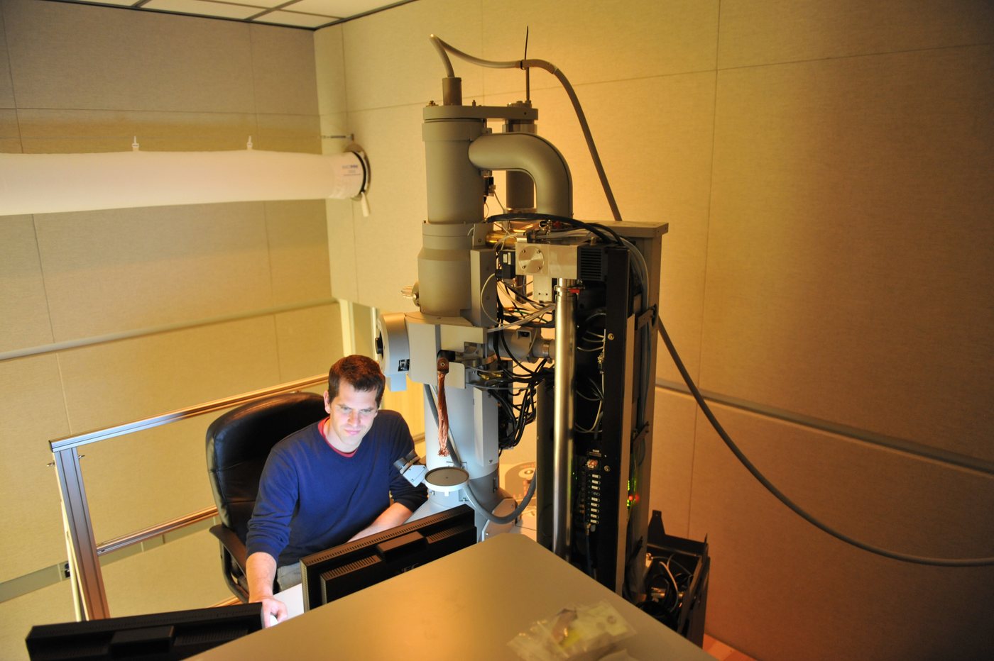A researcher seated at a tall grey transmission electron microscope column
                in a warmly lit room, surrounded by cabling and custom hardware.